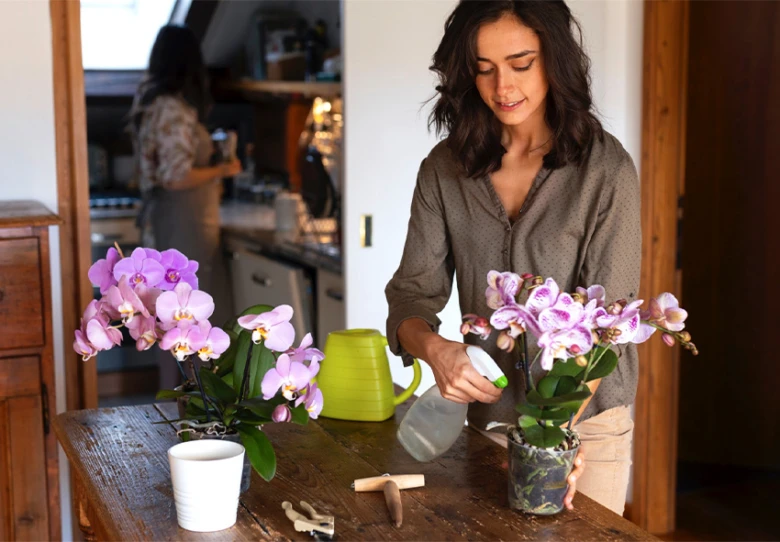 Žena zavlažuje fialovou orchidej na dřevěném stole s nástroji.